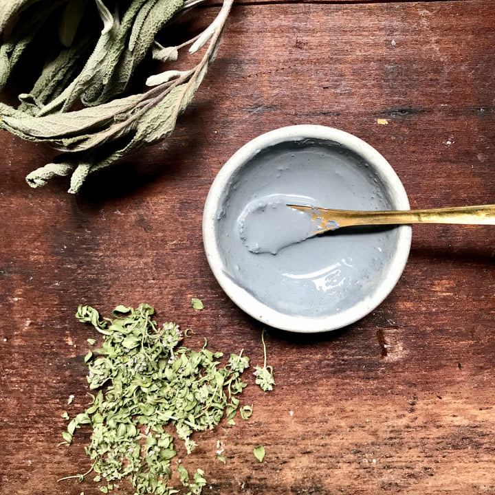 prepared facial mask in a bowl with a spoon. dried herbs in background.